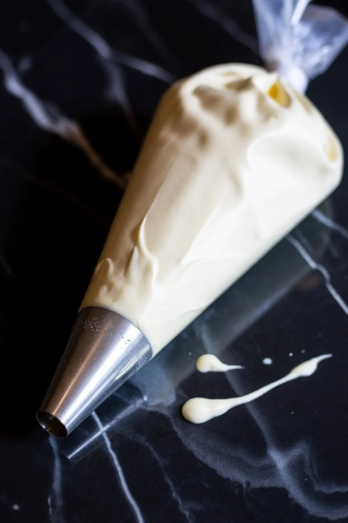 A piping bag filled with creamy white whipped topping rests on a dark marble surface with small drips beside the metal tip.
