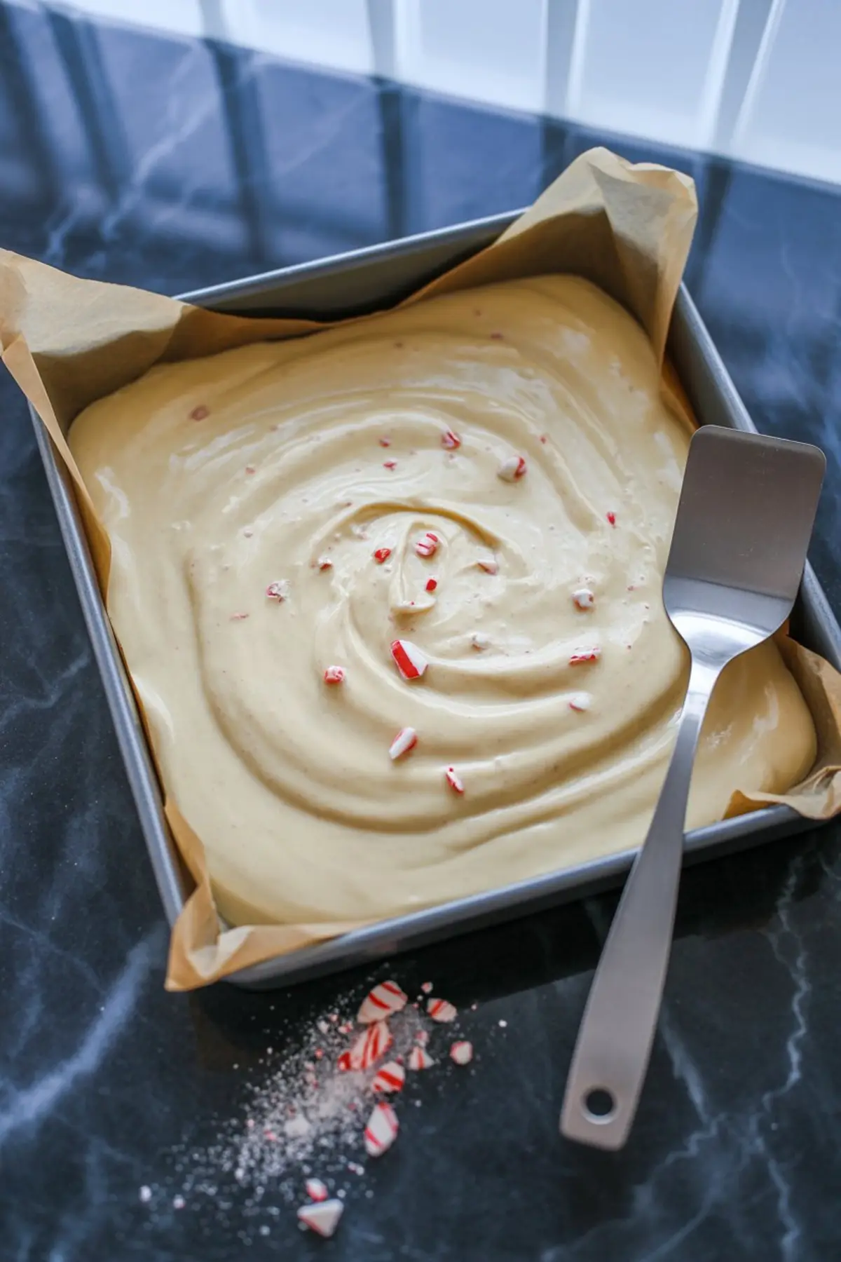 Square baking pan filled with smooth white chocolate fudge batter topped with crushed peppermint candy, resting on parchment paper with a metal spatula and peppermint crumbs on black marble surface.
