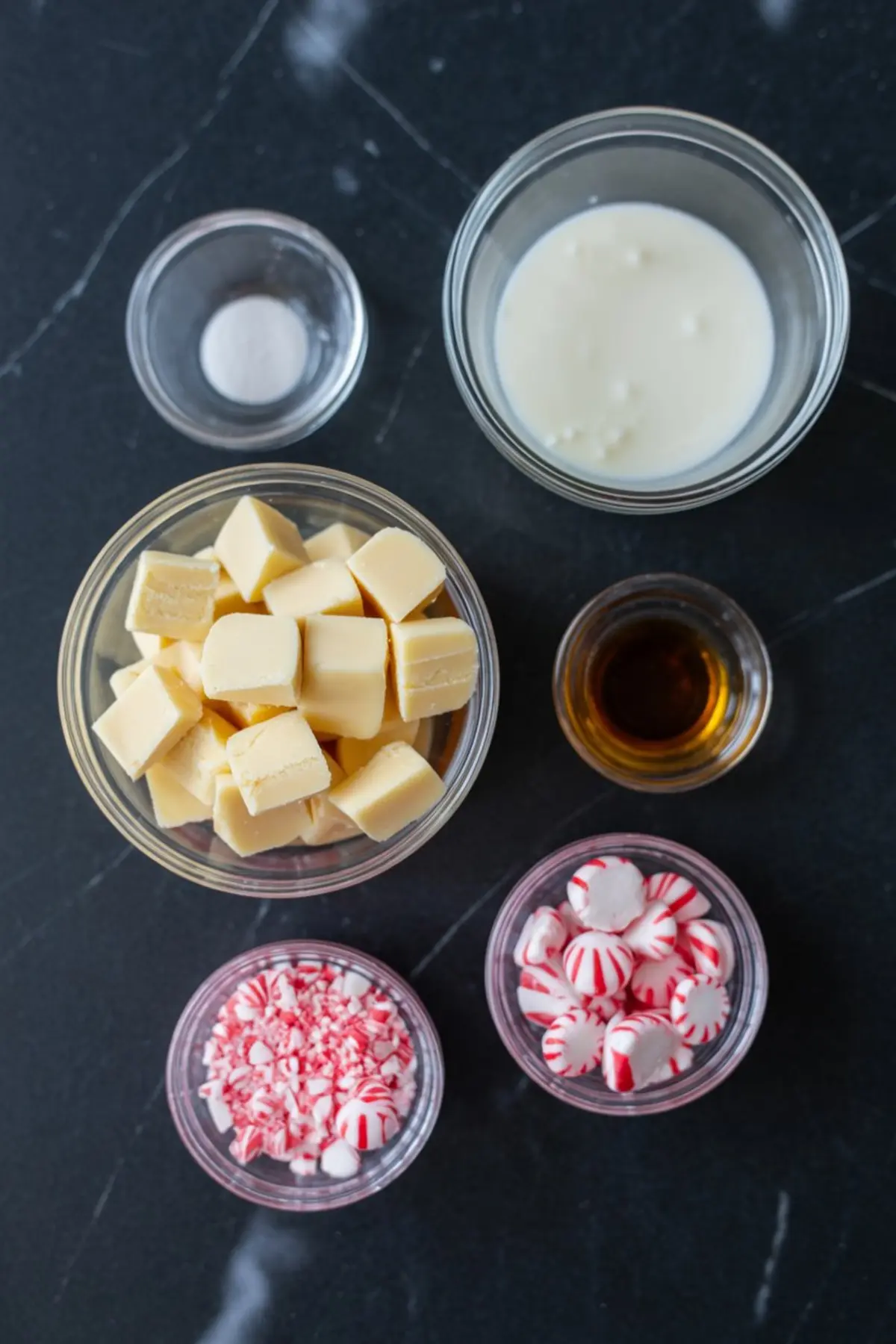 Overhead shot of fudge ingredients on a dark countertop, including white chocolate chunks, crushed peppermint candies, whole peppermint discs, sweetened condensed milk, vanilla extract, and baking soda in clear bowls.
