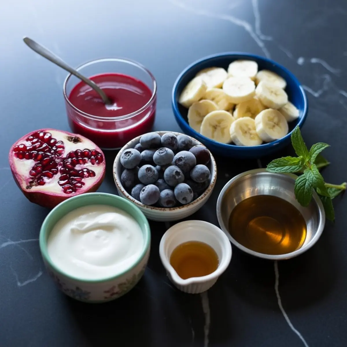 Flat lay of smoothie ingredients on a dark countertop, including a halved pomegranate, fresh blueberries, sliced bananas, Greek yogurt, honey, maple syrup, pomegranate juice, and mint leaves arranged in small bowls.