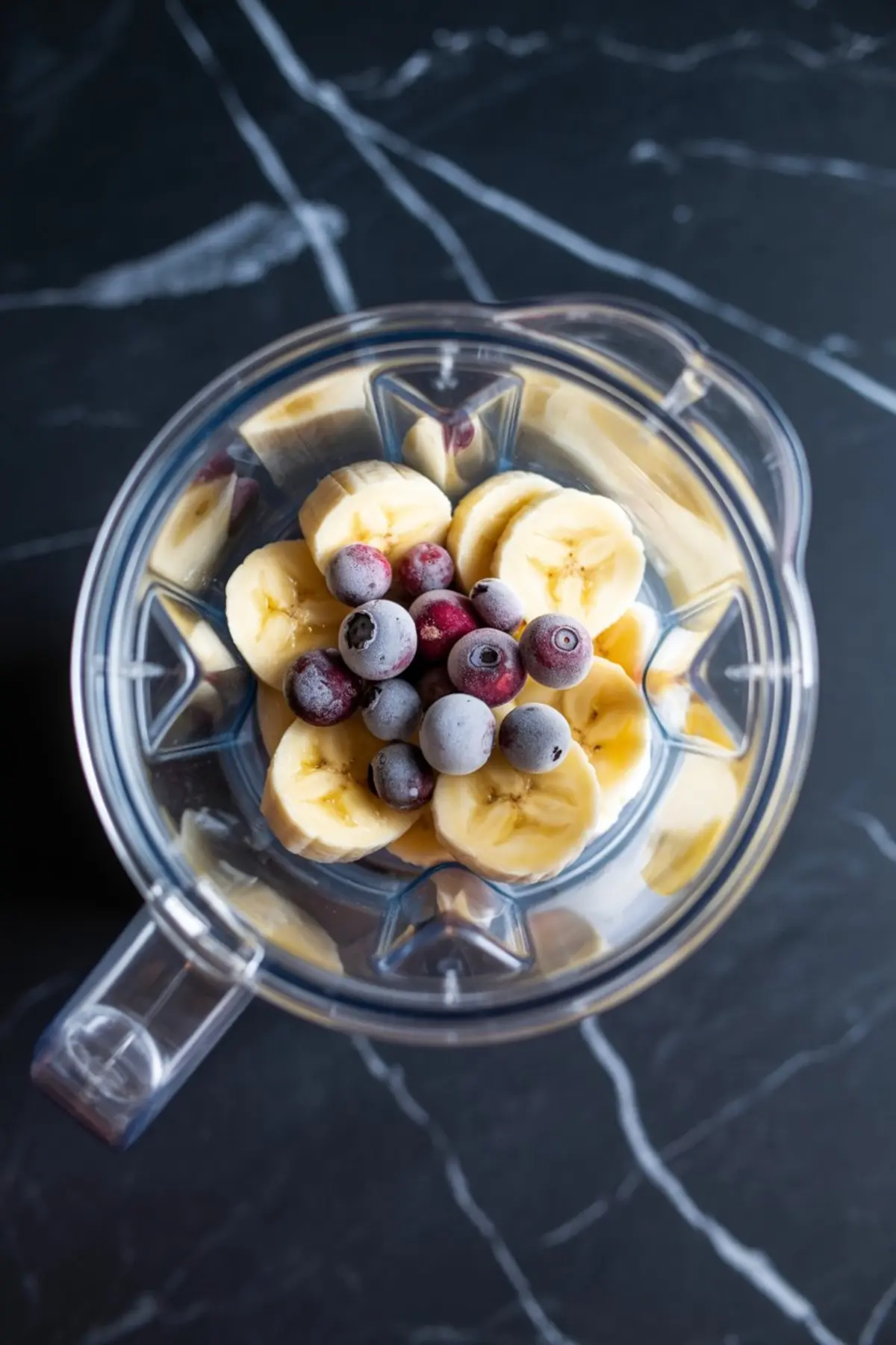 Top view of a blender filled with frozen blueberries and banana slices, prepared as the fruit base for a smoothie recipe on a black marble countertop.
