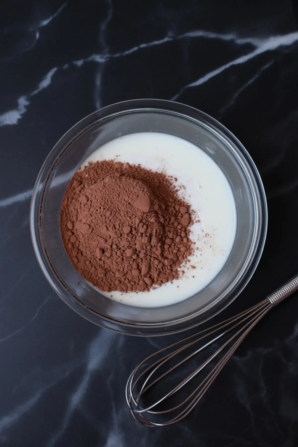 Mixing bowl filled with milk and cocoa powder with a metal whisk resting beside it on a dark marble background.