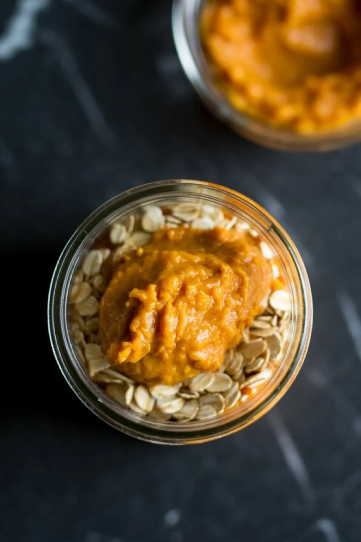 A glass jar filled with rolled oats topped with a generous scoop of pumpkin puree, captured on a dark marble surface for a moody autumn aesthetic.