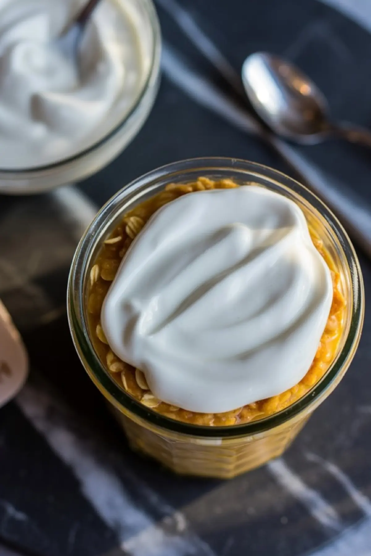 A jar of pumpkin overnight oats layered with oats and pumpkin puree, finished with a thick swirl of coconut cream or yogurt, styled with a spoon and bowl in the background.