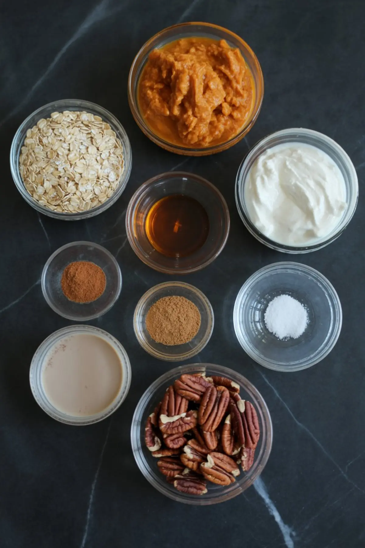 Flat lay of ingredients for pumpkin overnight oats, including rolled oats, pumpkin puree, Greek yogurt, maple syrup, cinnamon, nutmeg, salt, almond milk, and pecans arranged on a black surface.