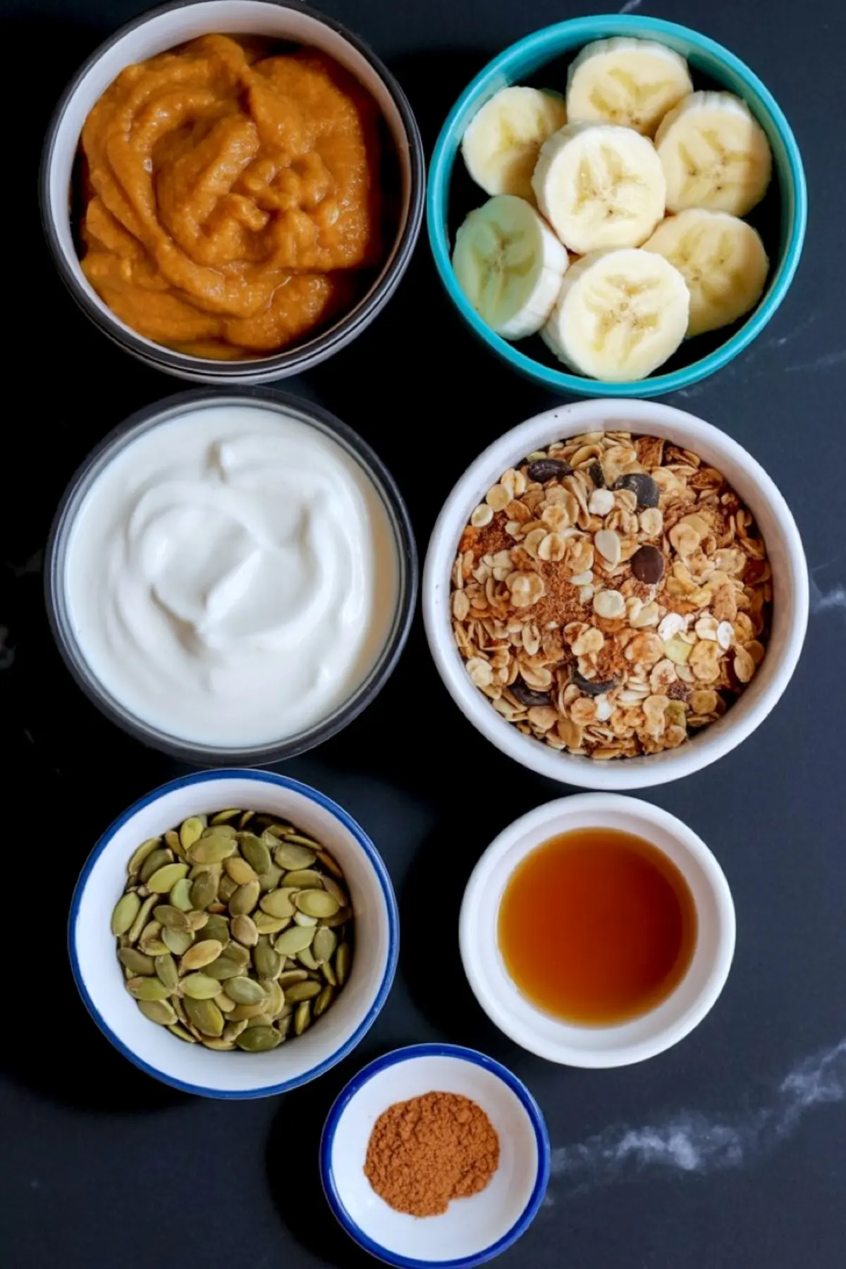 Flat lay of pumpkin pie smoothie bowl ingredients including sliced banana, pumpkin puree, granola, Greek yogurt, pumpkin seeds, maple syrup, and ground cinnamon in small bowls.

