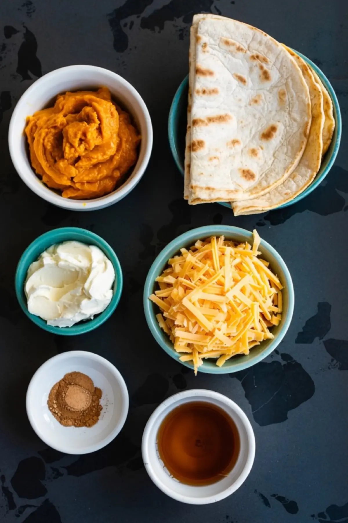 Flat lay of pumpkin spice breakfast quesadilla ingredients in small bowls, including pumpkin puree, shredded cheddar cheese, cream cheese, flour tortillas, ground cinnamon, and maple syrup on a dark surface.