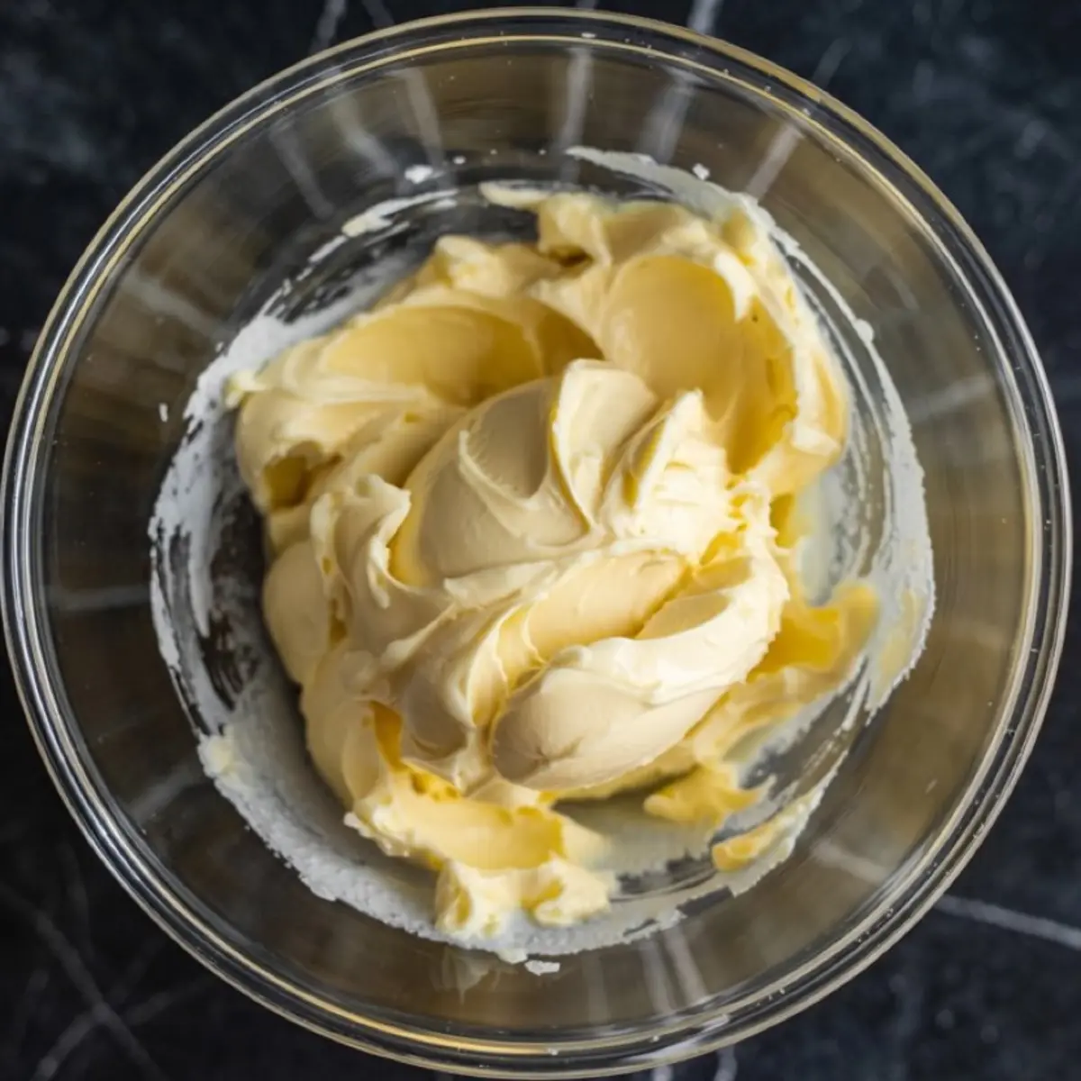 Creamy whipped butter in a clear glass mixing bowl, showcasing its smooth and fluffy texture on a dark kitchen surface.