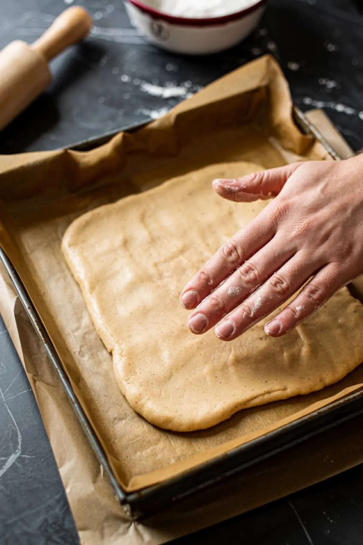 Hand pressing snickerdoodle dough evenly into a parchment-lined baking tray, preparing the base layer for bar cookies.
