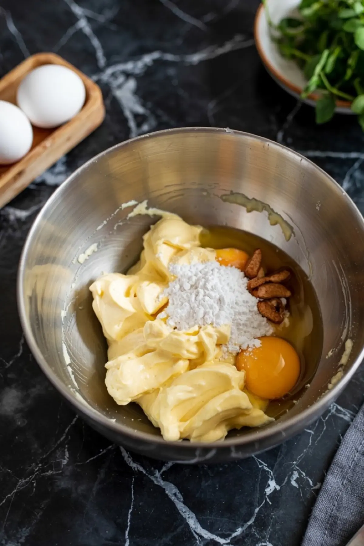 Butter, eggs, powdered sugar, and pecans in a metal mixing bowl on a black marble countertop, ready for mixing into snickerdoodle bar batter.
