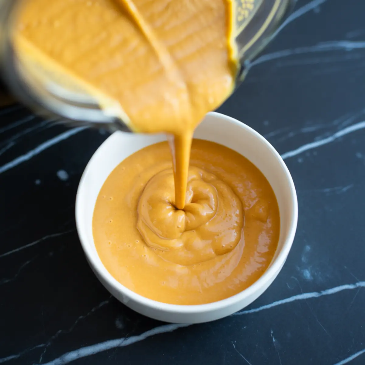 Creamy sweet potato smoothie being poured into a white bowl on a black marble surface, showcasing its thick, velvety texture and rich orange color.