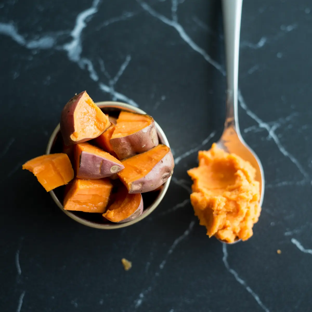 Cooked sweet potato chunks with skin in a small bowl next to a spoonful of mashed sweet potato on a dark marble surface.