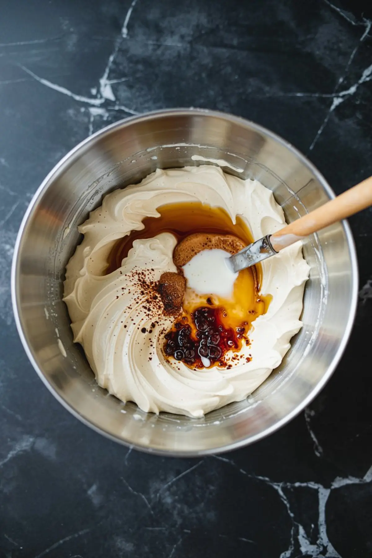 Whipped vegan frosting base topped with maple syrup, cinnamon, nutmeg, and non-dairy milk in a metal bowl, ready to be blended.