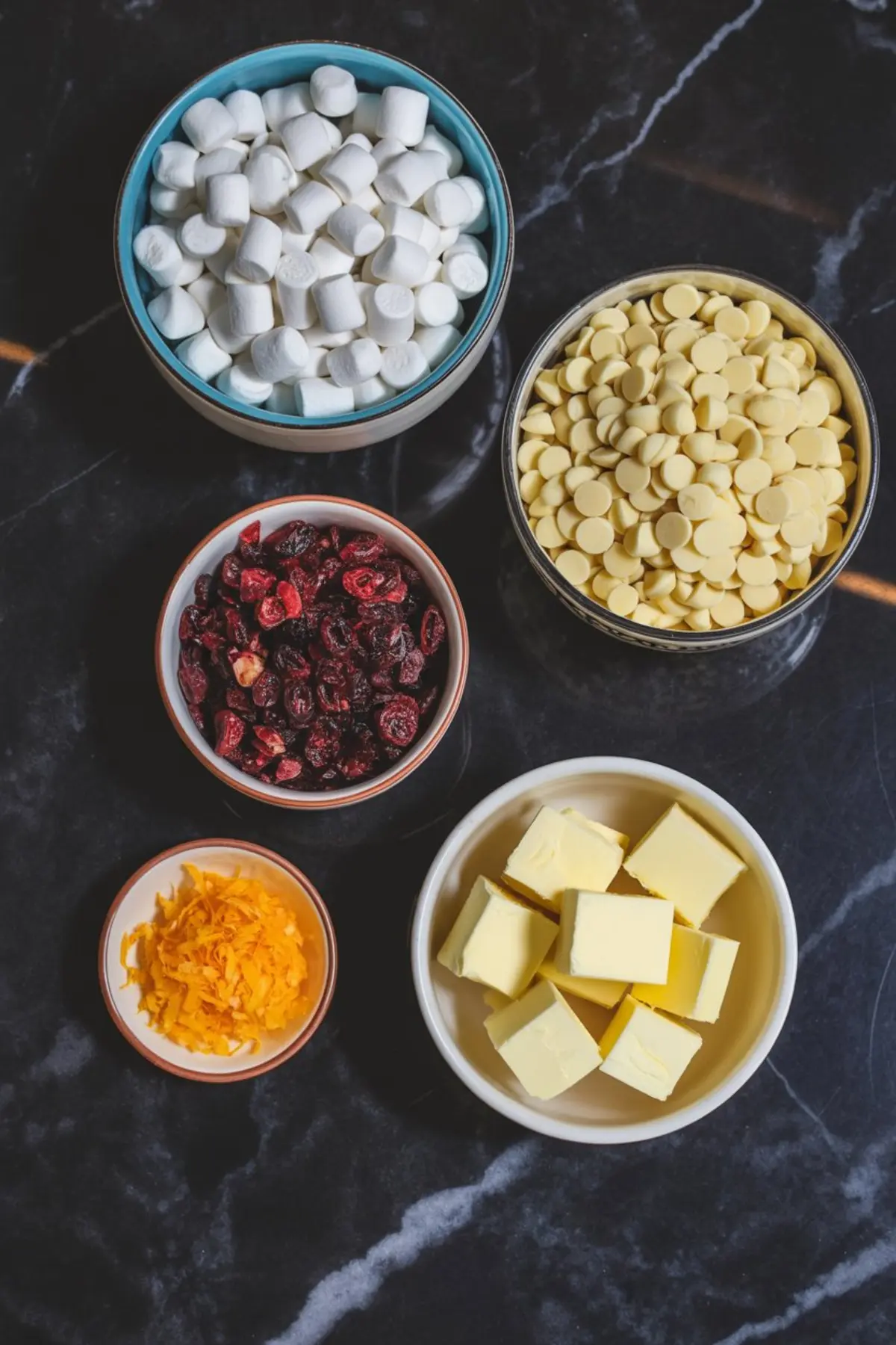 Flat lay of ingredients in bowls for white chocolate cranberry Rice Krispie treats, including mini marshmallows, white chocolate chips, dried cranberries, cubed butter, and fresh orange zest.