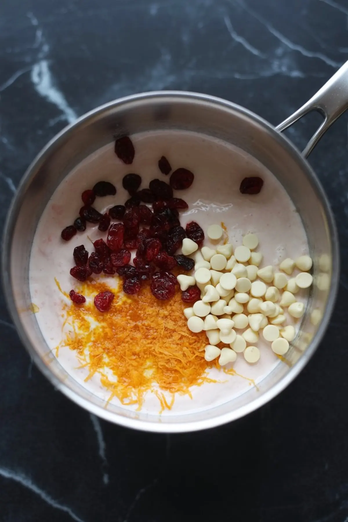 A saucepan filled with creamy marshmallow mixture topped with dried cranberries, white chocolate chips, and fresh orange zest on a black marble surface.