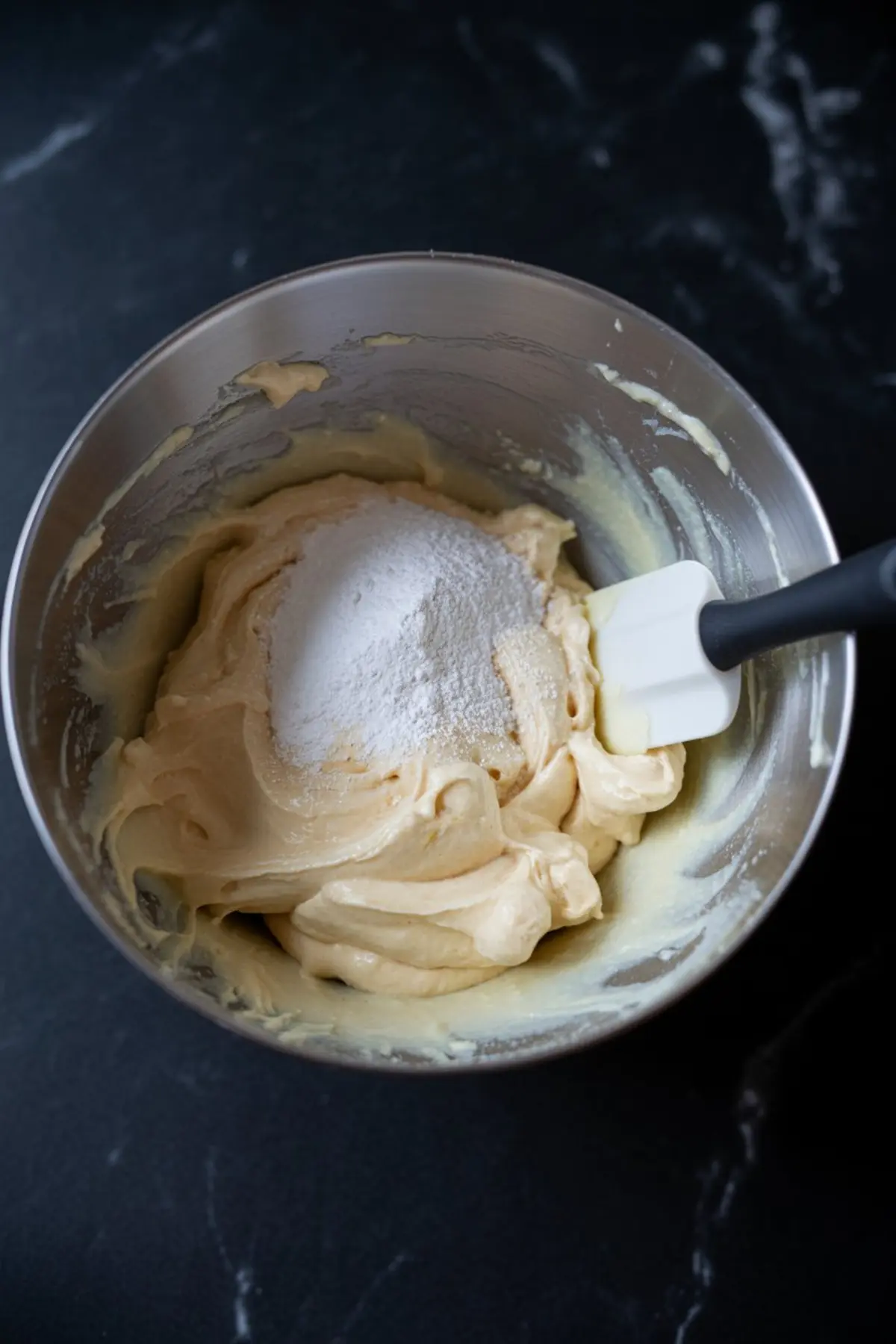 Bowl of mascarpone mixture being prepared with a spatula, featuring a heap of powdered sugar on top of a thick yellow cream base in a stainless steel mixing bowl.

