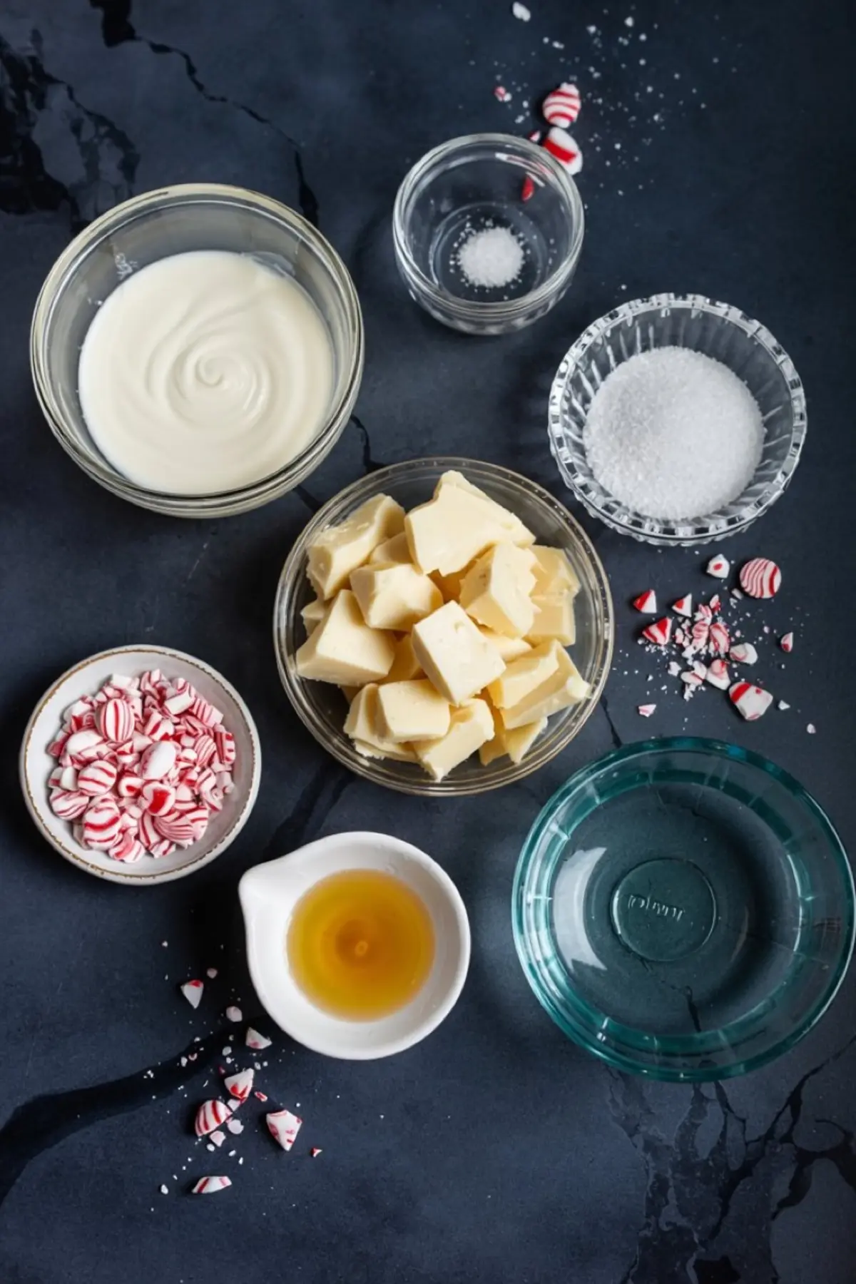 Flat lay of peppermint panna cotta ingredients on a dark marble background, including white chocolate chunks, crushed peppermint candies, granulated sugar, sea salt, vanilla extract, cream, and a glass of water.
