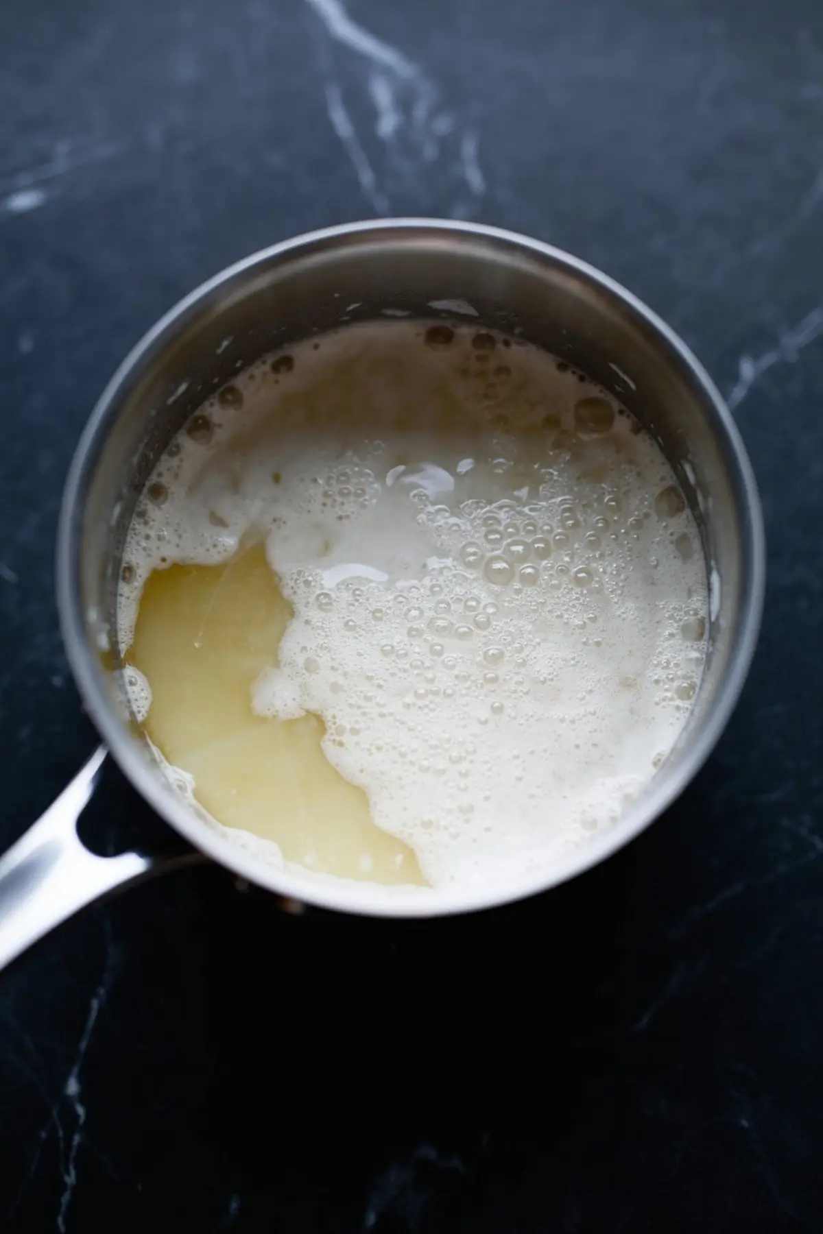 Overhead view of a saucepan containing a frothy cream and butter mixture on a black marble countertop, in the initial stage of preparing chocolate ganache.