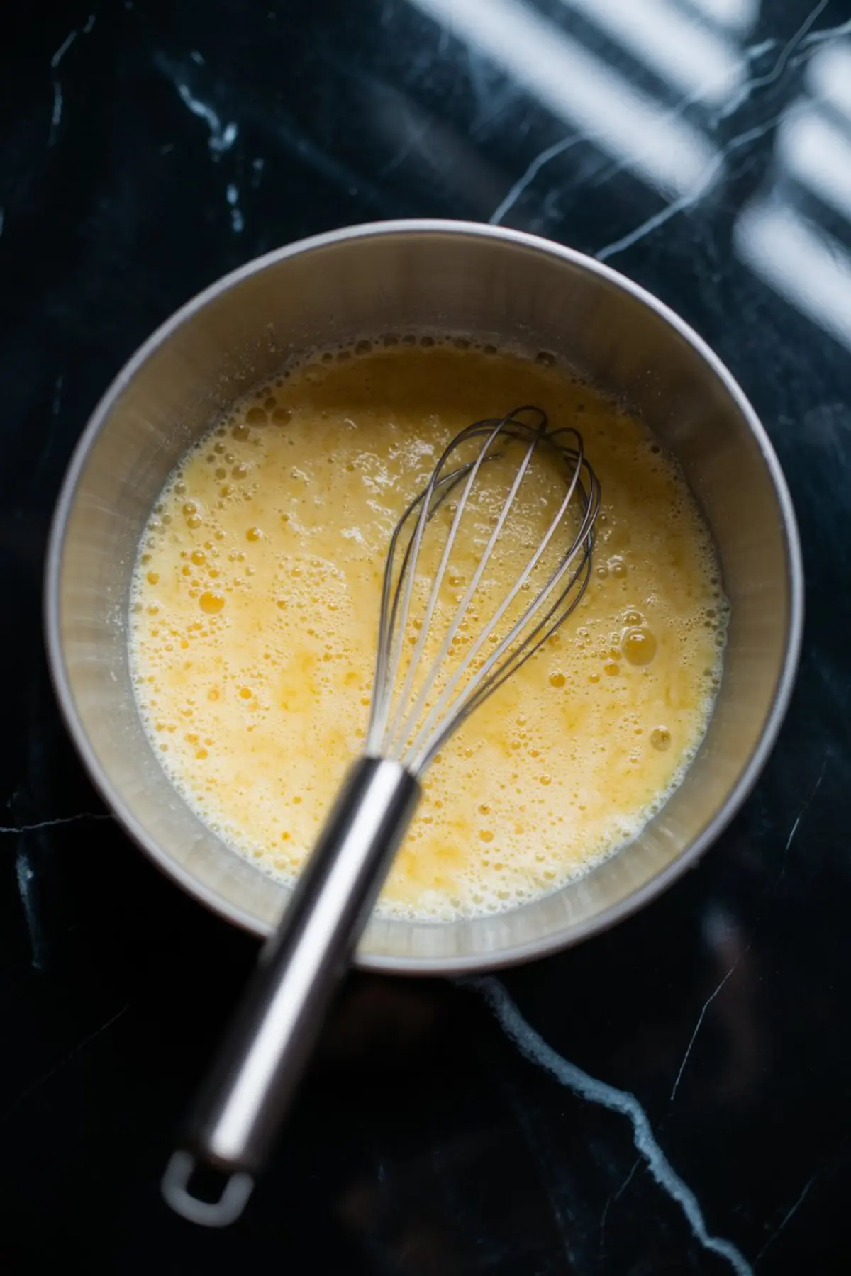 Beaten egg mixture with visible bubbles in a stainless steel bowl, with a metal whisk resting inside, prepared for tempering into the warm chocolate base.