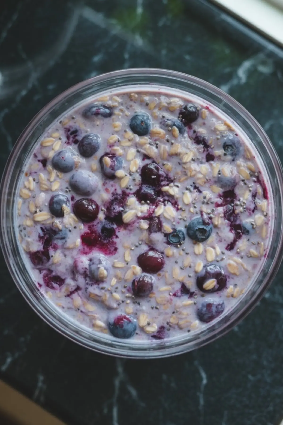 Top view of a creamy blueberry overnight oats mixture with visible rolled oats, whole blueberries, and chia seeds in a glass bowl on a dark marble countertop.