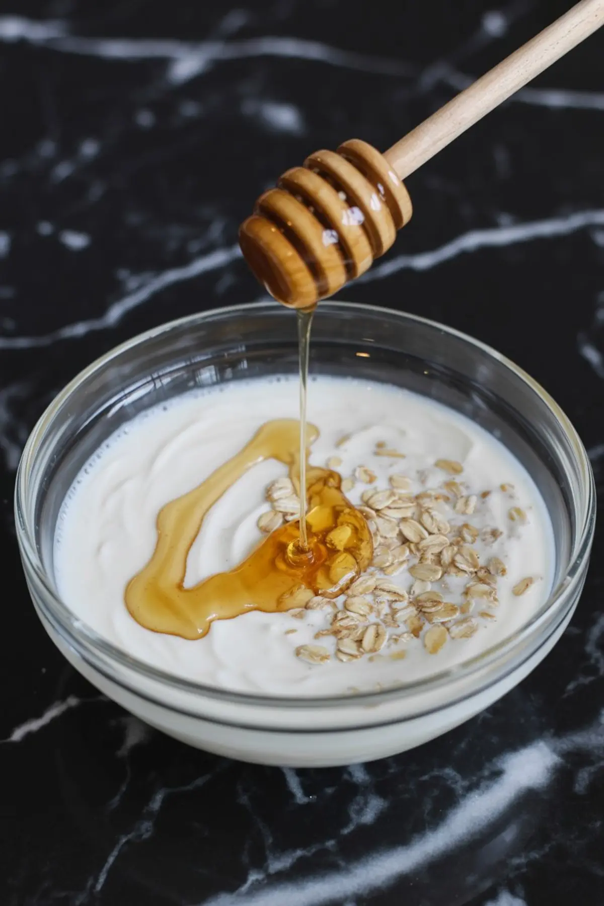 Honey being drizzled over a bowl of thick yogurt topped with raw rolled oats, set on a black marble surface for a healthy breakfast scene.