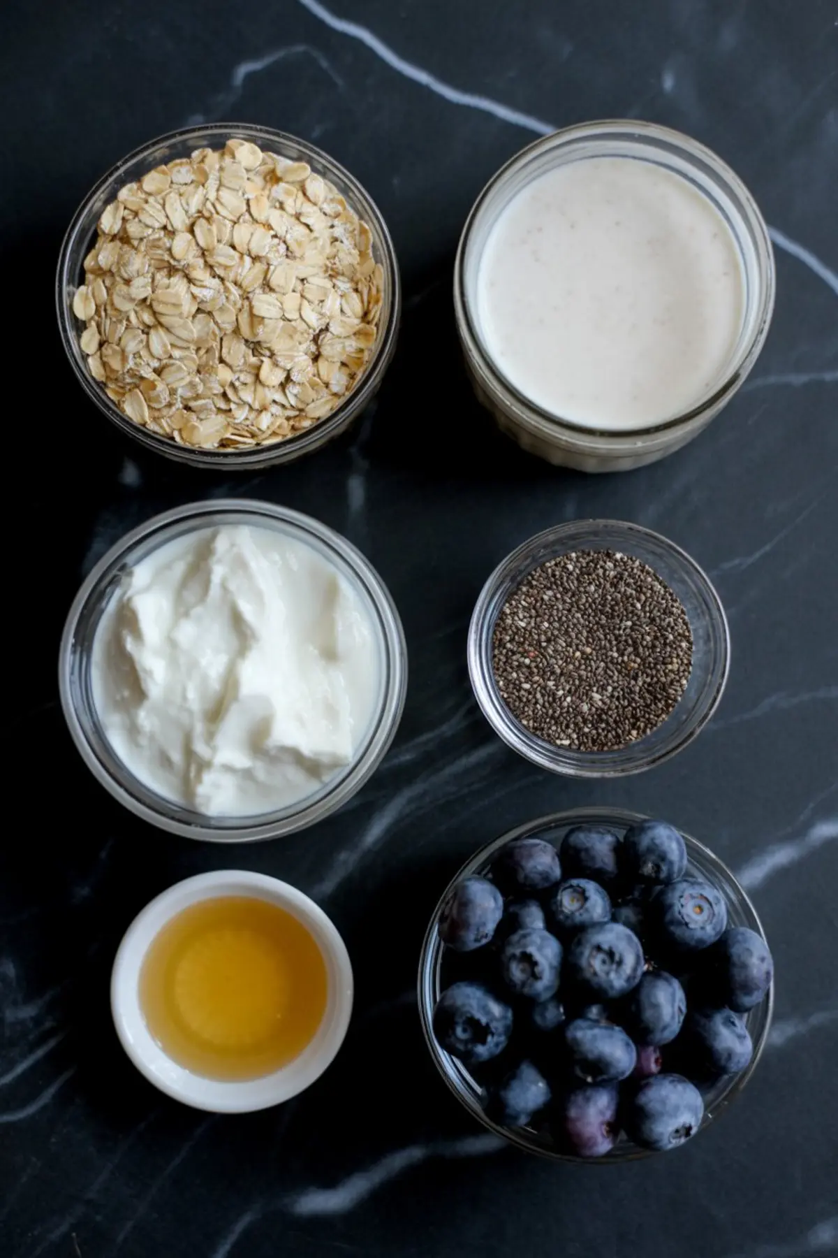 Flat lay of overnight oats ingredients including rolled oats, plant-based milk, Greek yogurt, chia seeds, honey, and fresh blueberries arranged in small glass bowls on a dark stone surface.