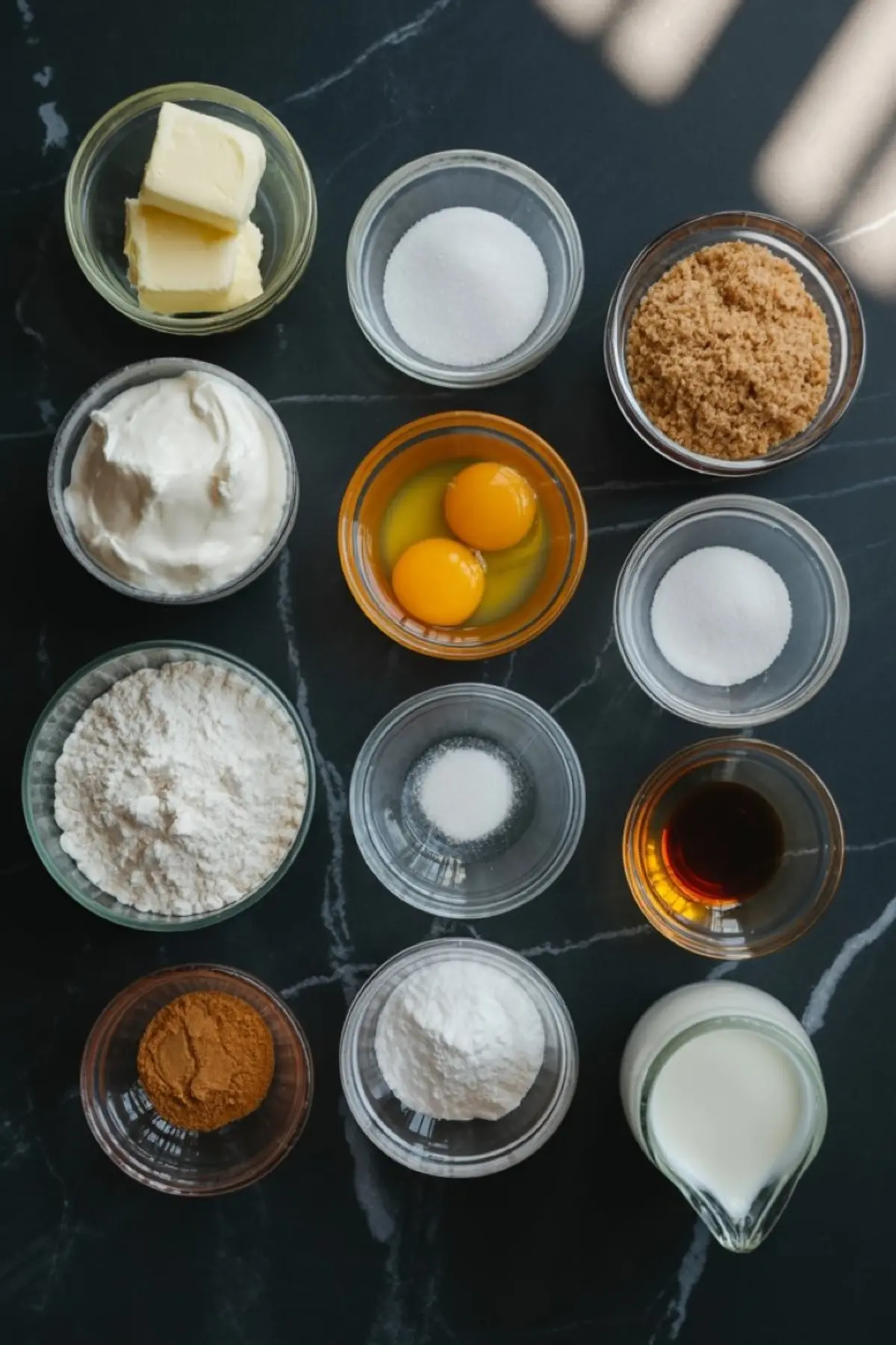 Overhead view of baking ingredients in glass bowls, including butter, sugar, brown sugar, eggs, flour, baking powder, cinnamon, yogurt, vanilla, and milk on a dark kitchen counter.