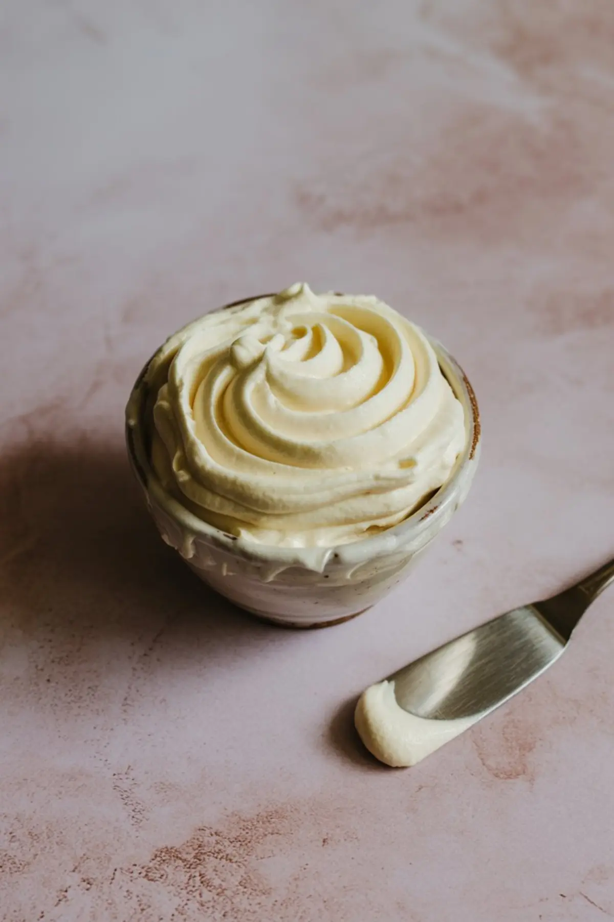 Swirled vanilla buttercream frosting in a small ceramic bowl with a spatula smear on a pink countertop, ideal for topping brownies or cupcakes.