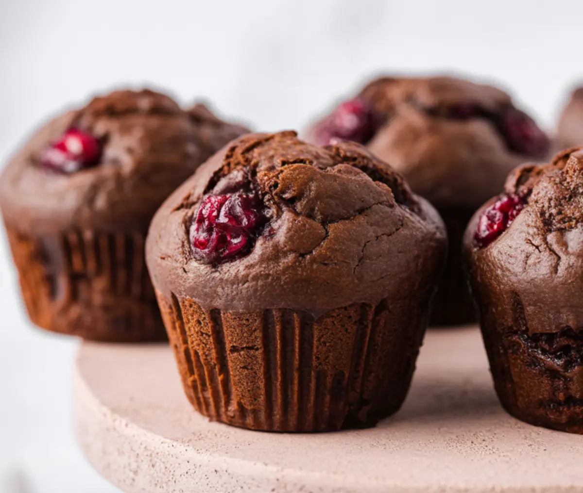 Chocolate cranberry muffins sit on a light stone stand. Soft chocolate crumb surrounds glossy red cranberries that peek through the tops. Warm brown tones highlight the rich batter and tender texture.
