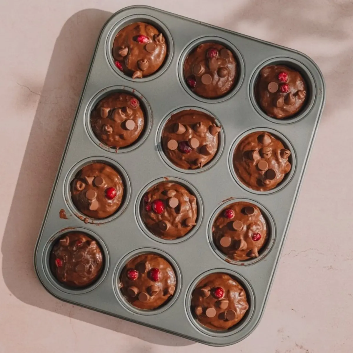 A metal muffin tin sits on a pale counter. Each cup holds chocolate muffin batter with chocolate chips and red cranberries scattered on top.