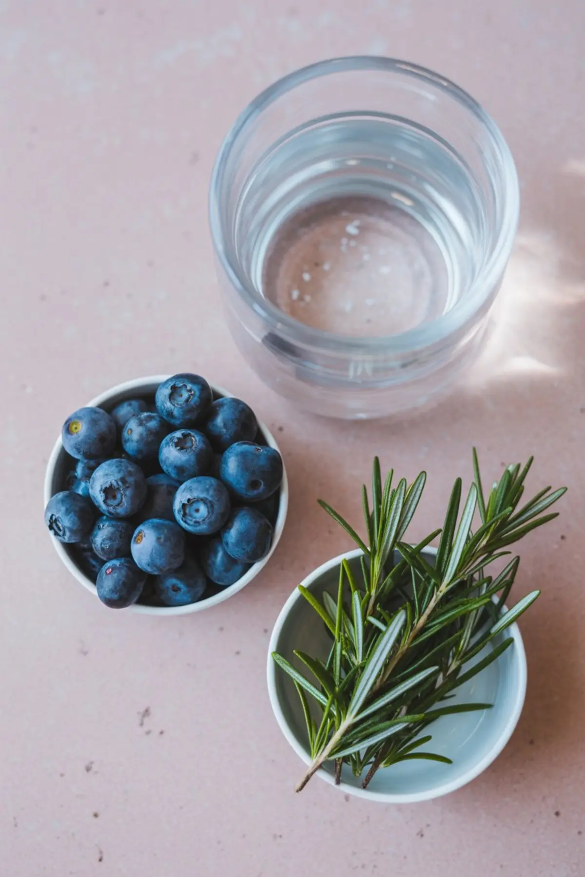 Flat lay of ingredients on a blush pink surface, featuring a bowl of fresh blueberries, a small bowl of rosemary sprigs, and a clear glass of water. The layout is simple and styled for a festive ice cube DIY.