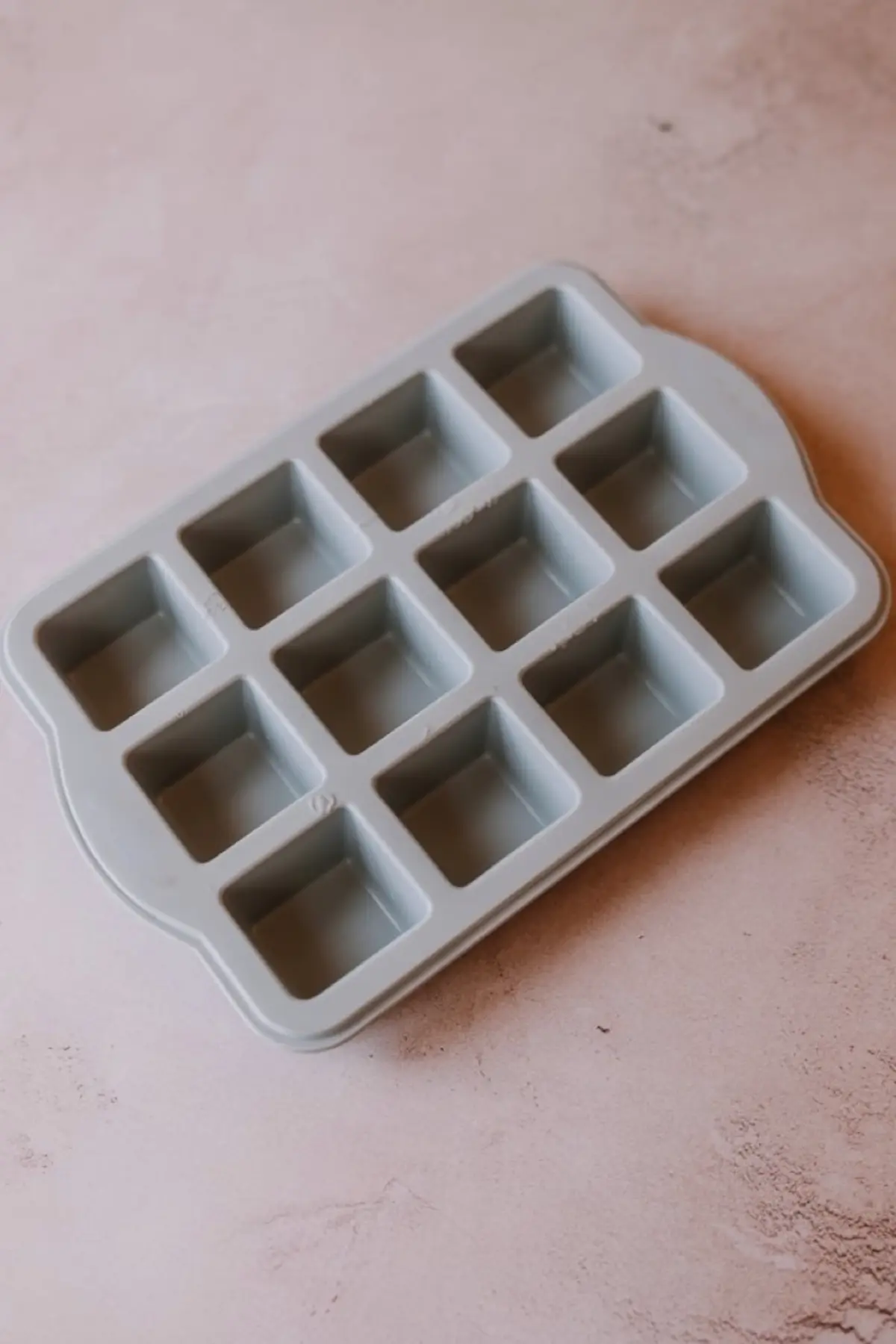 Empty gray silicone ice cube tray with twelve square compartments placed on a soft pink textured background, ready for filling with ingredients.