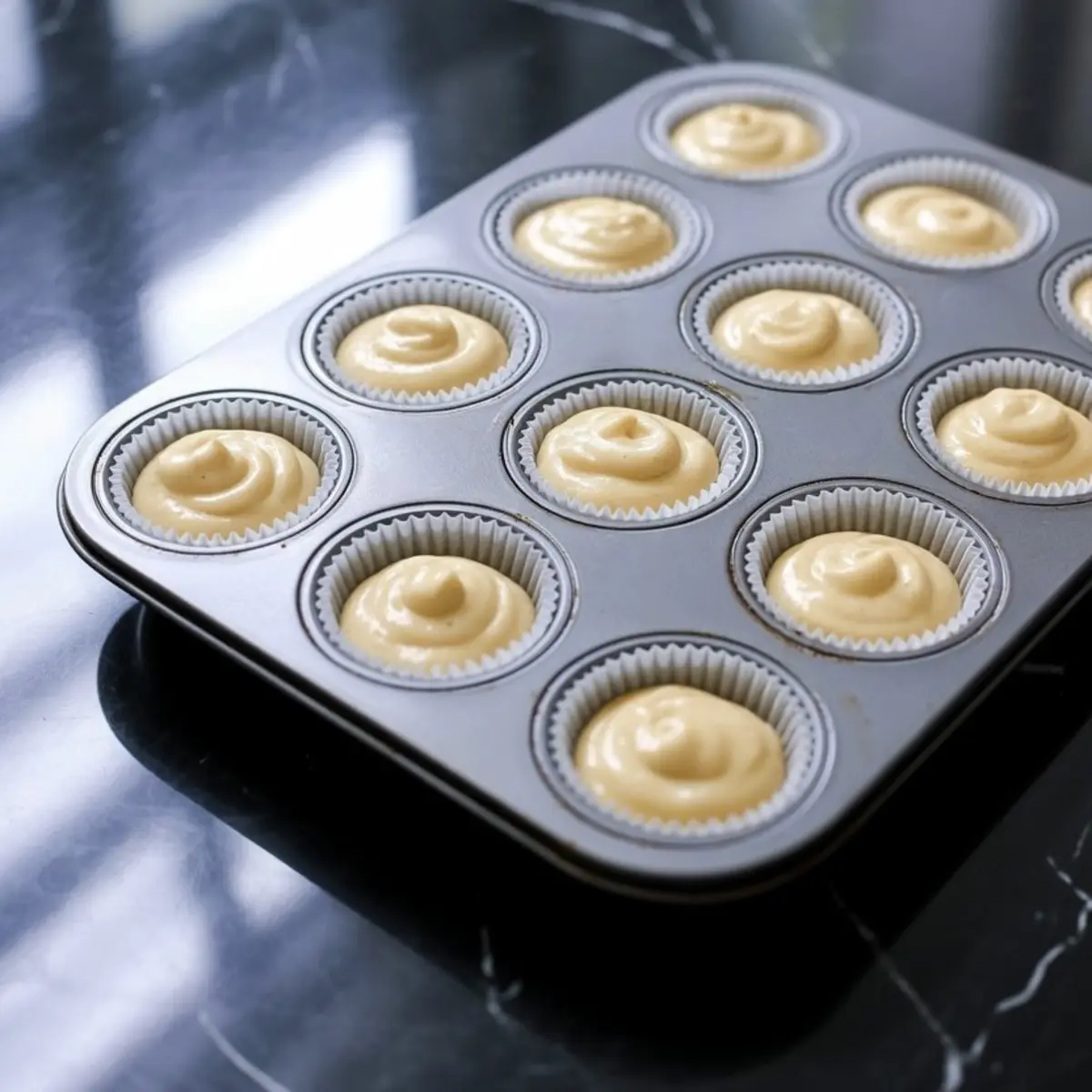 Cupcake batter evenly distributed in white paper liners inside a metal muffin tray, ready to be baked on a glossy black counter.