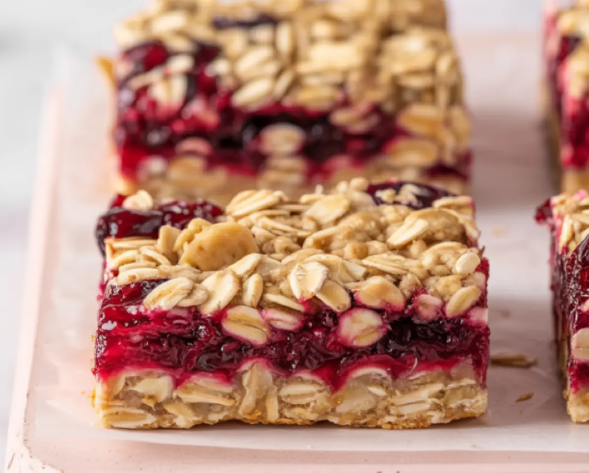 Close-up of a cranberry oatmeal bar with a thick oat crust, red cranberry filling, and oat crumble topping on a pink surface.