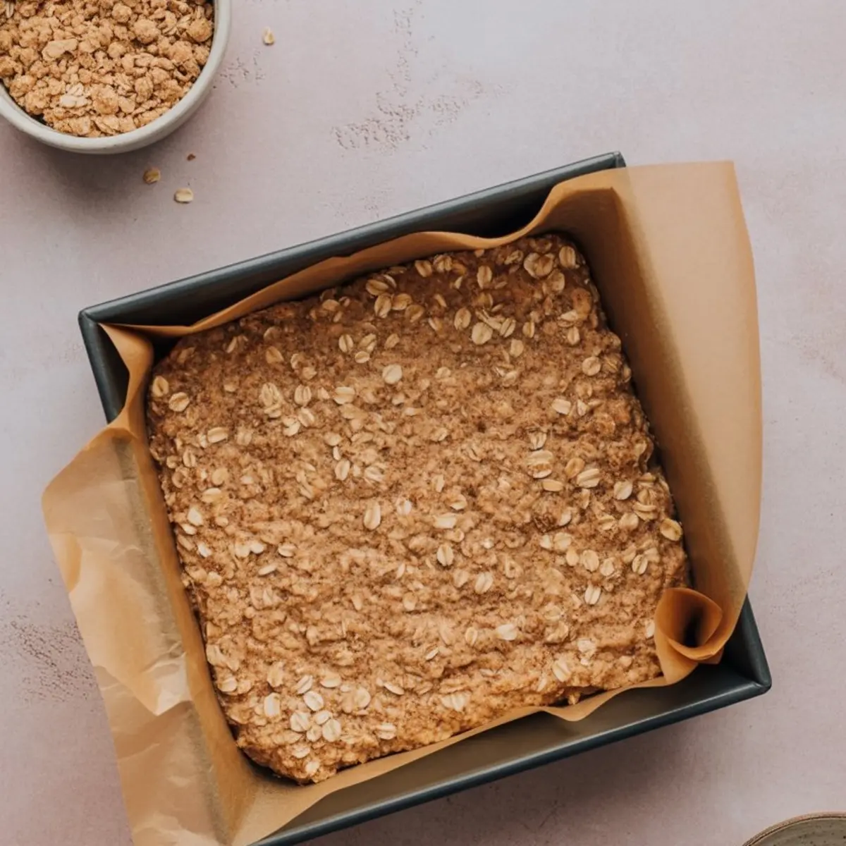 Baking pan lined with parchment paper holding an unbaked oatmeal crust mixture topped with oats.