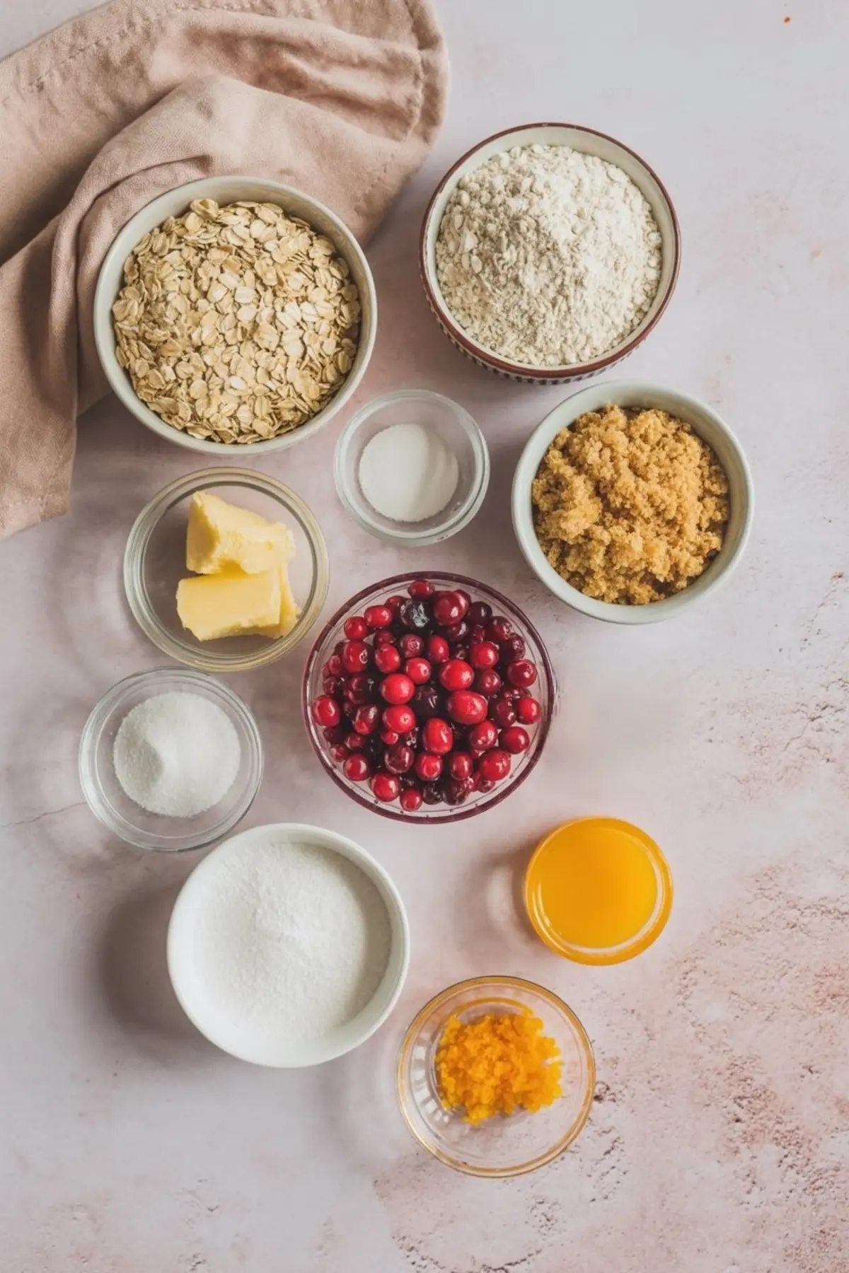 Flat lay of baking ingredients for cranberry oatmeal bars including oats, flour, brown sugar, cranberries, butter, sugar, orange zest, and juice in small bowls.