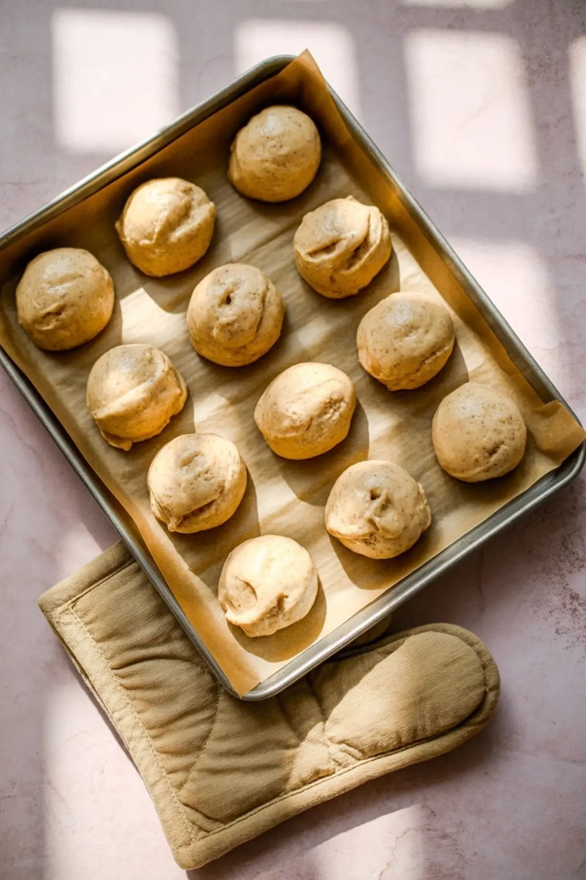 Unbaked scoops of eggnog cookie dough arranged on a parchment-lined baking tray, lit with natural light and paired with a tan oven mitt, showcasing the dough’s spiced texture before baking.

