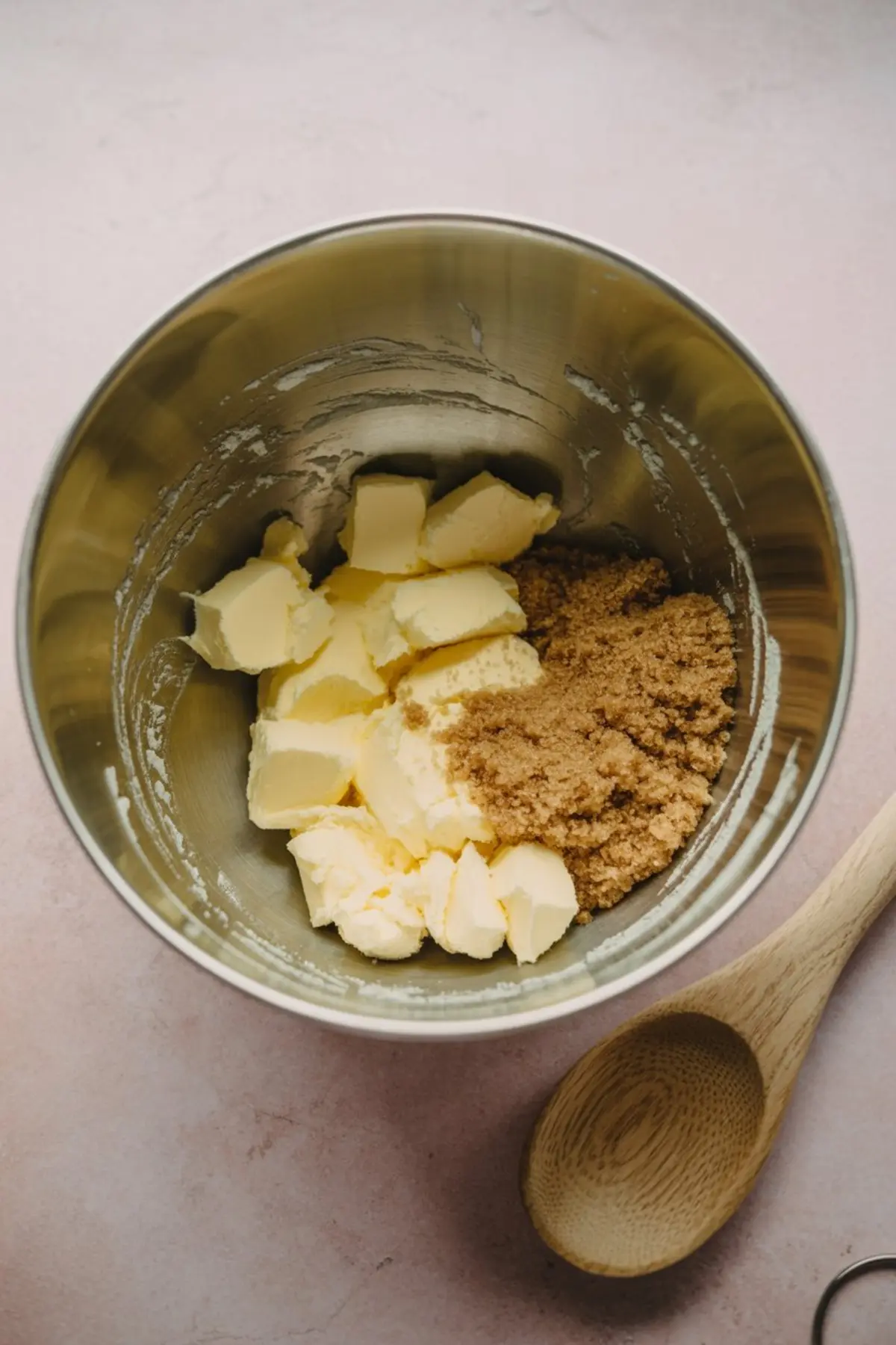 Cubed butter and brown sugar combined in a stainless steel mixing bowl with a wooden spoon on the side, showing the early stage of creaming ingredients for holiday cookies.
