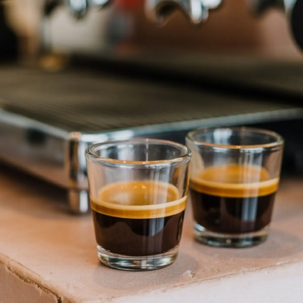 Two espresso shots with golden crema served in clear glass cups, placed in front of a stainless steel espresso machine.
