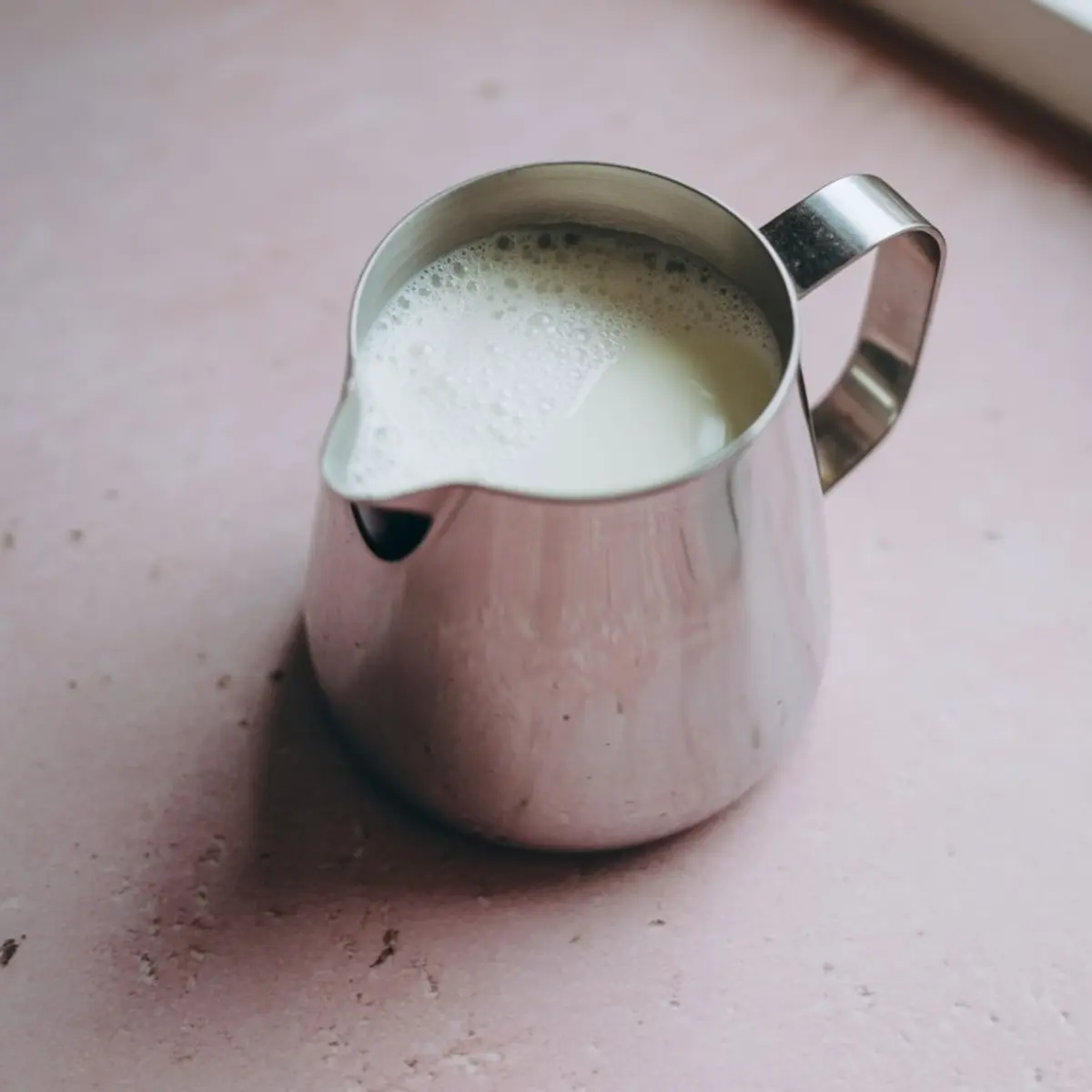 Stainless steel milk frothing pitcher filled with warm, foamy milk, placed on a pastel pink countertop.
