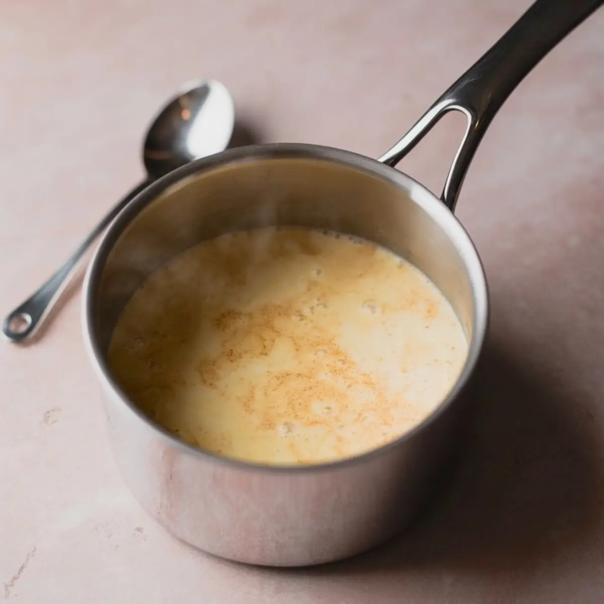 Steaming saucepan of eggnog mixture with cinnamon specks, sitting on a soft pink surface beside a silver spoon.

