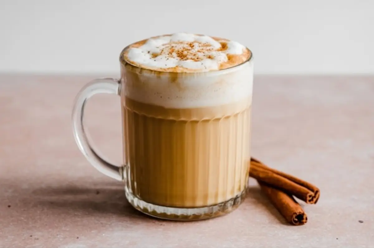 Eggnog latte in a ribbed glass mug topped with frothy cream and cinnamon, styled with cinnamon sticks on a pale pink surface.
