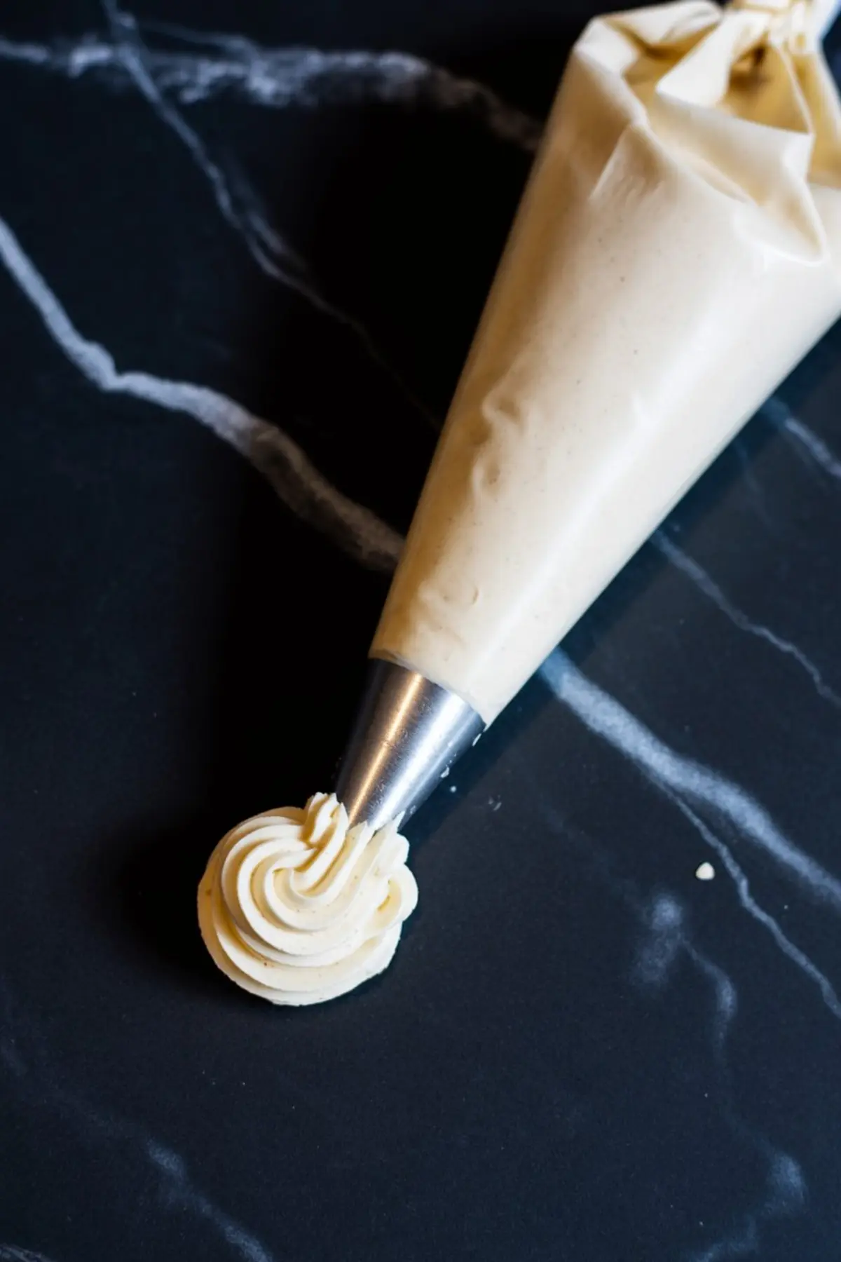 Piping bag filled with smooth, pale tan ermine frosting, forming a decorative swirl on a dark marble surface.
