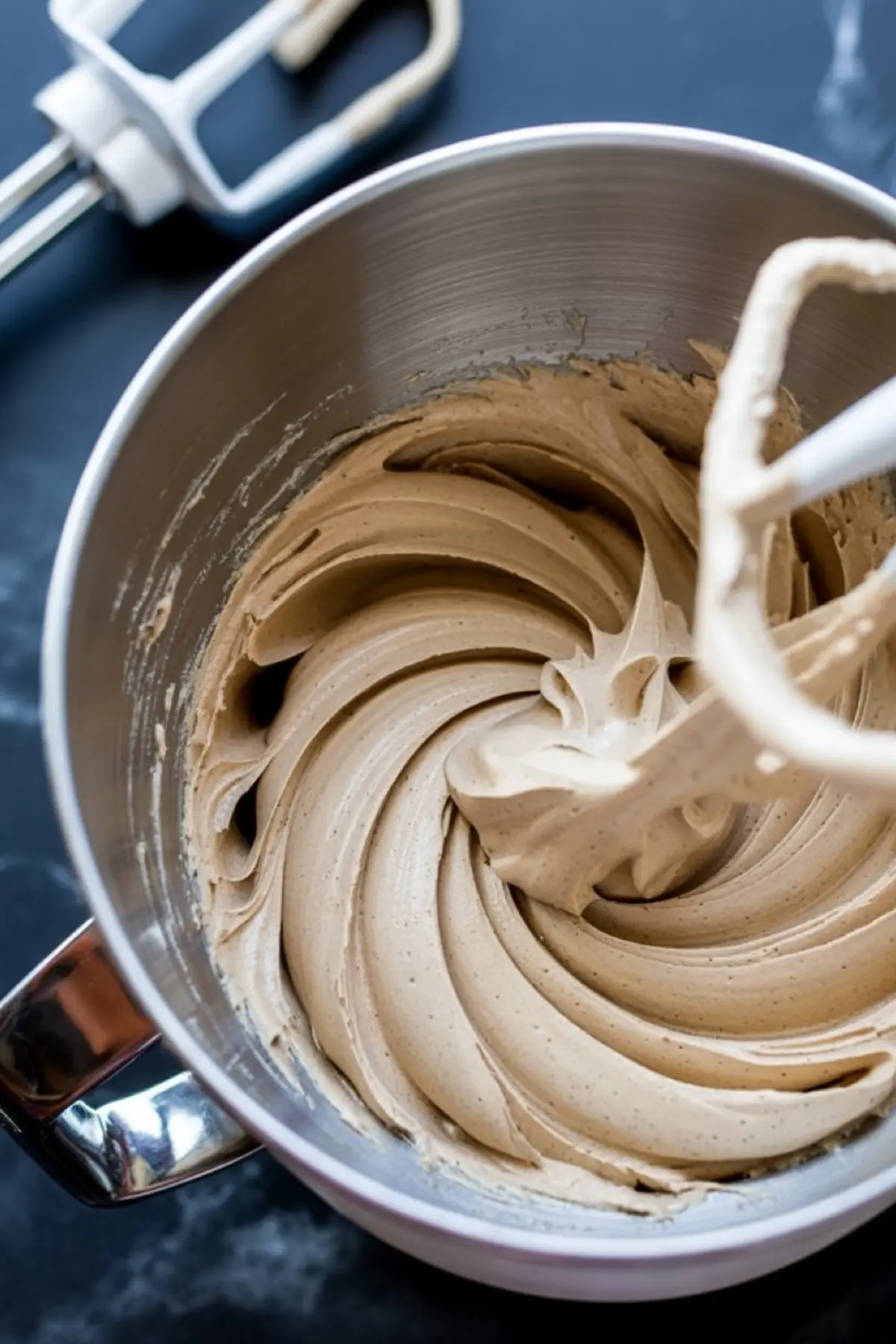 Whipped ermine buttercream frosting in a metal mixing bowl, forming soft peaks with a swirl pattern created by a paddle attachment.
