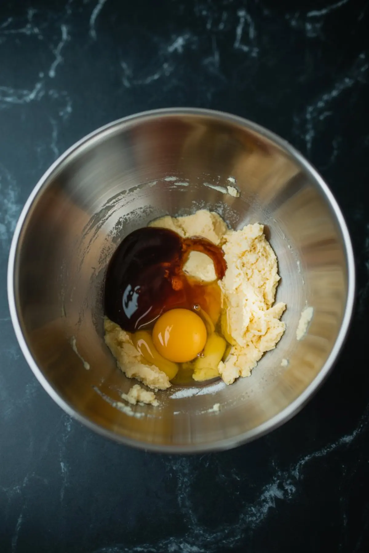 Egg and vanilla extract added to creamed butter and sugar in a metal mixing bowl, showing a step in cookie dough preparation.
