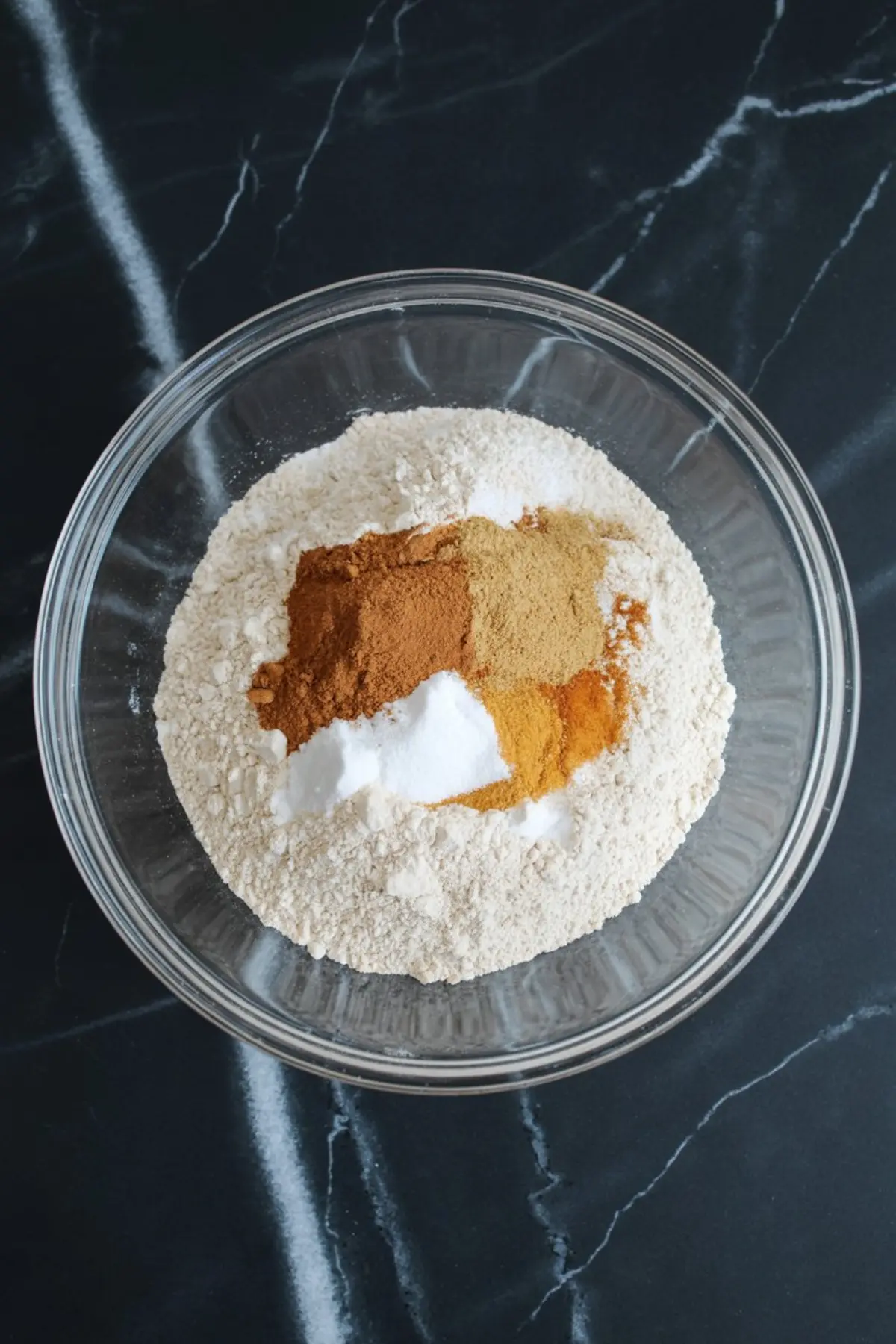 A glass bowl holds a blend of dry baking ingredients including flour, cinnamon, ginger, baking soda, and turmeric on a dark marble surface.
