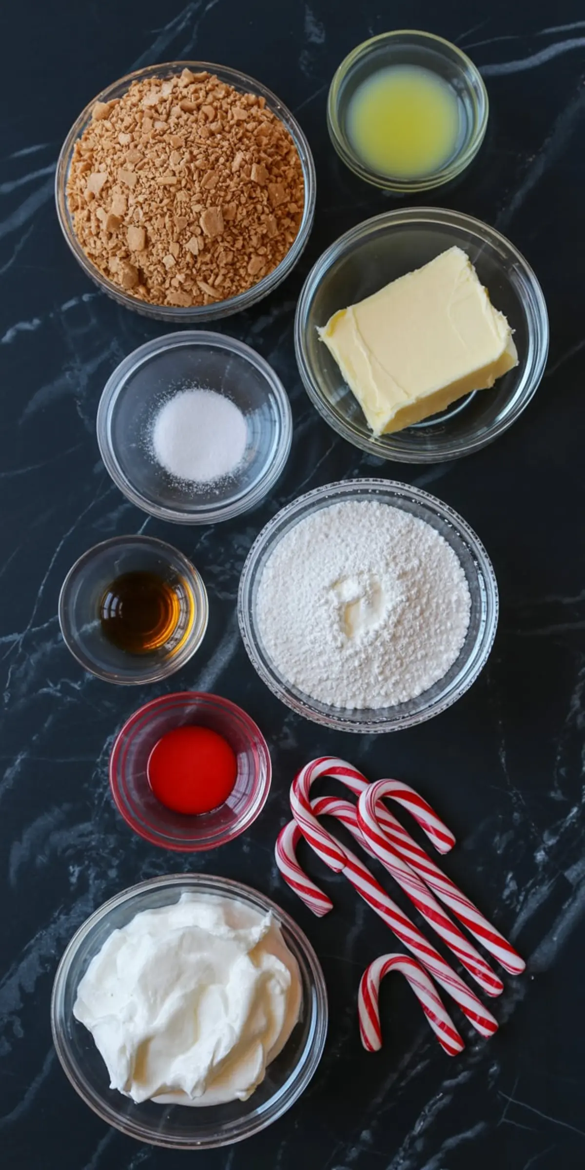 Flat lay of peppermint cheesecake mousse ingredients on black marble, featuring crushed cookies, butter, powdered sugar, whipped topping, peppermint extract, vanilla extract, candy canes, and melted butter in glass bowls.
