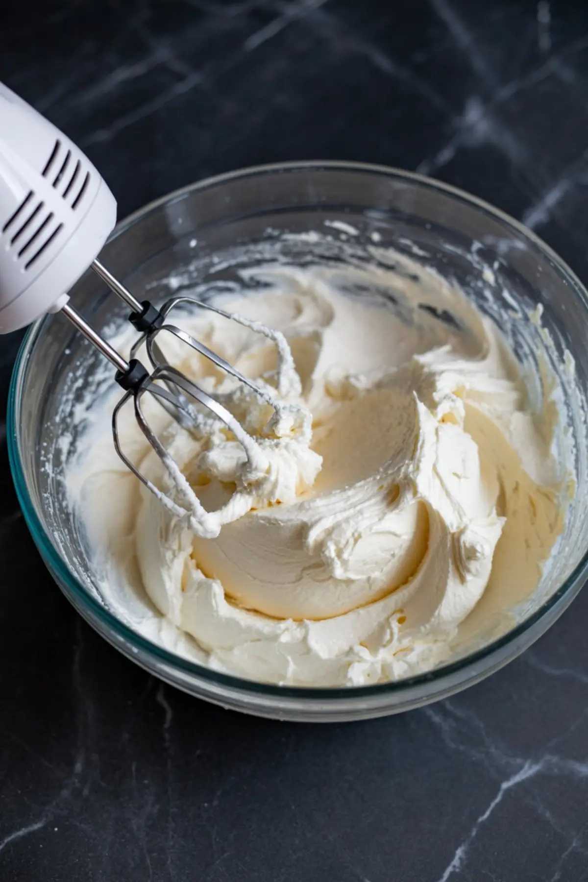 Electric hand mixer blending whipped cream and cream cheese mixture in a glass bowl, creating a smooth mousse base for peppermint cheesecake dessert.
