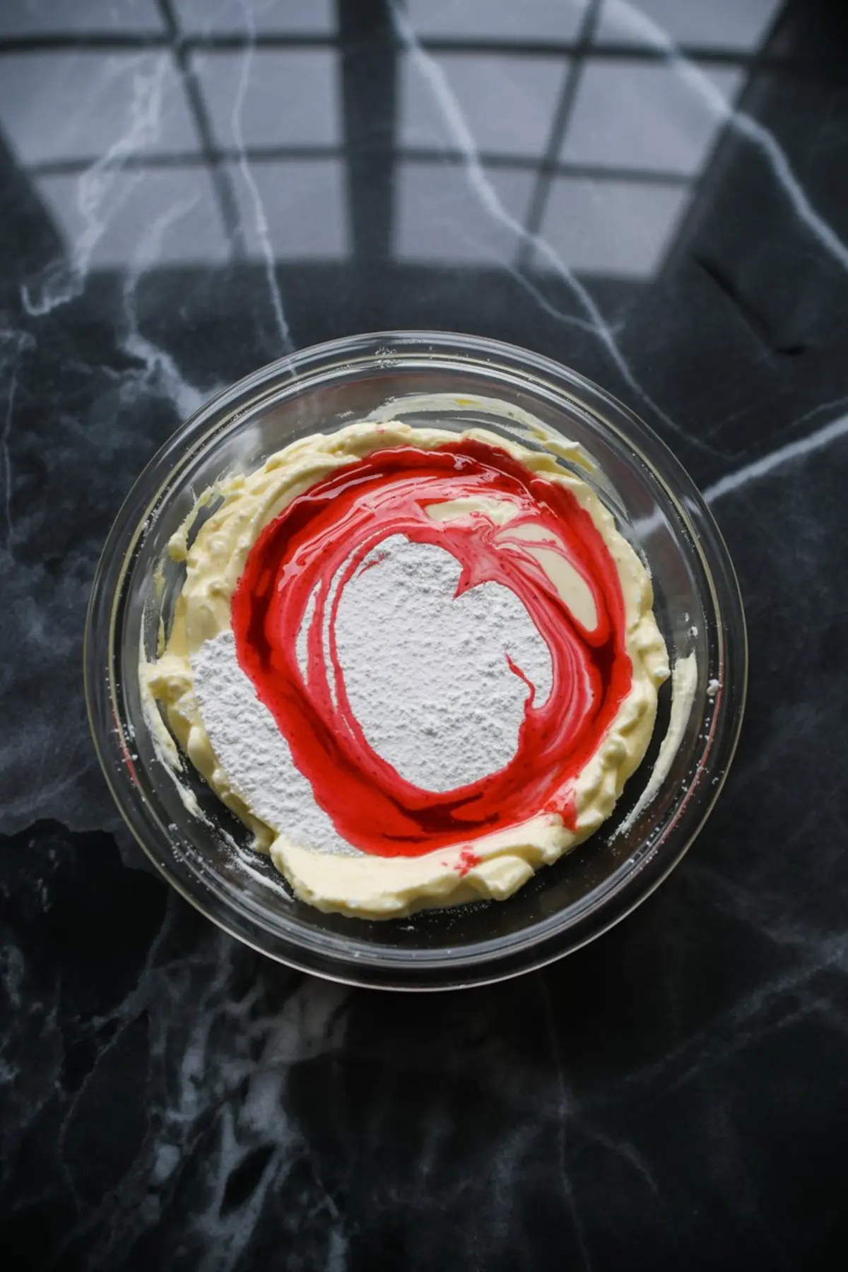 Glass mixing bowl filled with a creamy base, powdered sugar, and red peppermint extract swirled on top, ready to be mixed for a peppermint cheesecake mousse.
