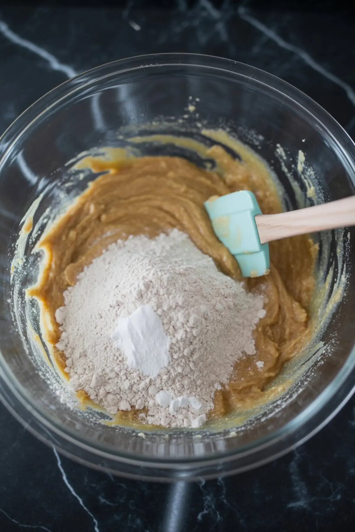 A mixing bowl holds cookie batter with a pile of flour and baking powder on top, next to a silicone spatula, showcasing the process of making blondie batter.
